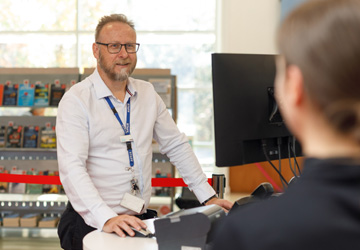 A library staff member stands facing a customer who is to the right of the frame